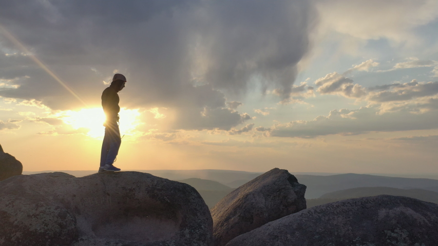 A young male climber walks along the top of a mountain range and jumps over a chasm at sunset.