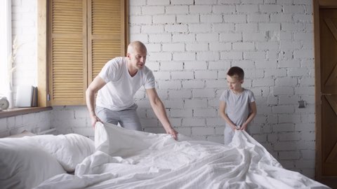 Slowmo shot of father giving five to son helping him make bed, then ruffling his hair with affection