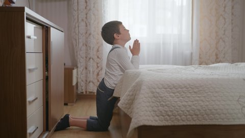 religion, little Christian boy with faith and hope in heart with closed eyes clasped hands prays to God on his knees in room near bed against window
