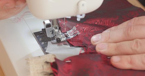 Handsome Male hand pushes red paisley lining material through a sewing machine during suit making process. Fashion, creation and tailoring. High angle view