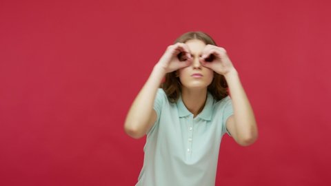 Nosy inquisitive young woman in t-shirt making glasses shape with hands, looking through fingers in binoculars gesture and expressing surprise, shocked by seen. studio shot isolated on red background