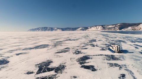 Khivus boat moves on frozen ice of lake Baikal on sunny winter day. Beautiful natural landscape of snow-capped mountains under blue sky and modern vehicle is moving along frost surface of siberian sea