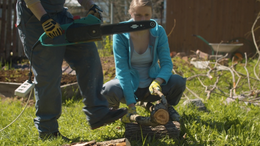 Two women working in the courtyard of country house. Mature cutting tree with an electric chain saw, young helping she. Active and healthy lifestyle concept