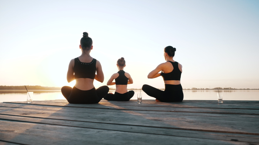 Diverse Group of Three Calm Female Synchronously Awe Do Yoga Exercise, Meditation Sitting in Lotus Position on Boardwalk Near Water Together Outdoor. Asana for Energy Balance, Mental Wellness, Harmony Royalty-Free Stock Footage #1052927954