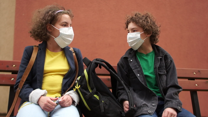 Back to school after quarantine. Cute curly boy and girl are sitting on a bench at a safe distance and talking. Children say goodbye contactlessly, touching their elbows Royalty-Free Stock Footage #1052931629