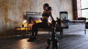 Young attractive african american woman blogger recording video with a smartphone live streaming doing sport exercises in bedroom at home. Quarantine workout concept
