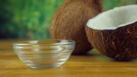 Pouring natural coconut oil into a glass bowl placed on a wooden platform. Closeup shot of a brown hairy coconut, a coconut shell and a bowl of organic coconut oil - blurred green background