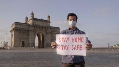 A young confident man or male wearing face mask standing and holding a placard with message 'Stay home stay safe' during city lockdown amid coronavirus or COVID 19 in front of empty Gateway of India. 
