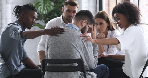 Young mixed race diverse compassionate people supporting desperate unhappy man. Depressed male patient suffering from bullying, sharing personal problems at psychological therapy group meeting.