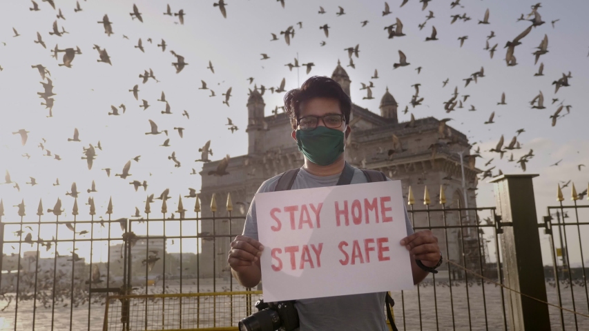 A young man wearing face mask standing and holding a placard with message 'Stay home stay safe' during city lockdown amid coronavirus in front of Gateway of India with pigeons flying in back. Royalty-Free Stock Footage #1052948441