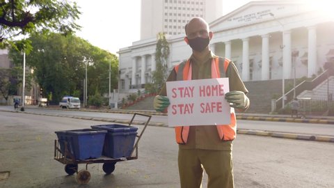 A male street sweeper essential service provider standing and holding a placard with message 'stat home stay safe' during city lockdown amid coronavirus or corona epidemic or pandemic