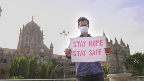 A young man wearing face mask standing and holding a placard with message 'Stay safe stay home' during city lockdown amid coronavirus or COVID19 in front of historic landmark CSTM VT terminus station
