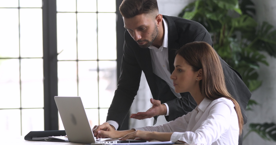 Side view confident company employee in suit standing near table, helping female colleague with corporate computer software. Young businessman discussing reviewing project with skilled manager. Royalty-Free Stock Footage #1052948801