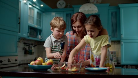Happy family of mother and two kids cooking apple pie together with eggs, sugar, flour and apples at home. Kid helping mom to slice apples and put into bowl on blue kitchen.  Arkistovideo