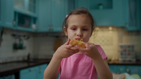Preschool girl in pink t-shirt eating homemade pizza standing on blue kitchen at home. Kid enjoy eating pizza with hands. 