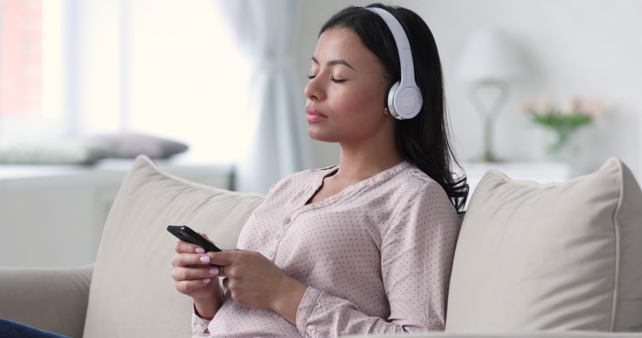 Peaceful african ethnicity young woman wearing headphones, listening affirmations, visualizing future alone on comfortable sofa. Happy tranquil woman listening slow pacifying music from mobile app. Royalty-Free Stock Footage #1052950559