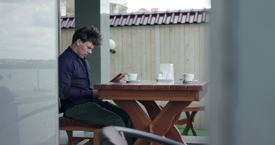 Worried businessman sitting in open cafe, thinking and typing on a tablet in windy day. Slowly in 4K