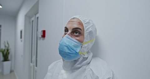 Tired female medical worker with face scars from protective uniform after overwork with patients coronavirus infected taking off protective mask and resting in corridor of modern clinic