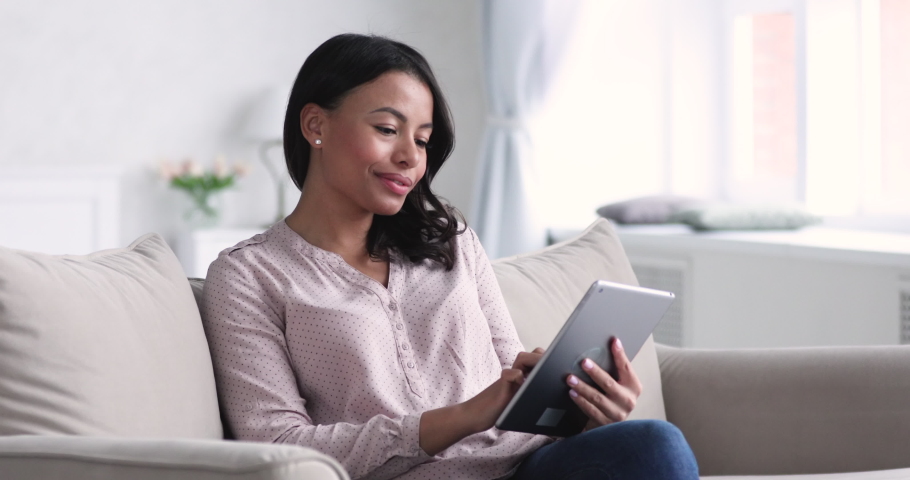 Beautiful young african american woman sitting om comfortable sofa, using digital computer tablet at home. Relaxed mixed race businesswoman web surfing information online, working alone from home. Royalty-Free Stock Footage #1052959559