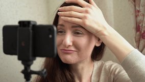 Beautiful Caucasian brunette woman holds selfie stick, looks at herself in screen of phone. Girl make faces, upset, sad, smile, scare because of skin. Female covers her wrinkles with hand. Take photo