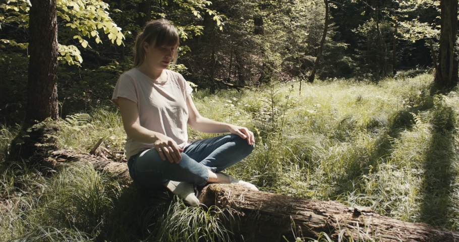 Woman practicing yoga meditation sitting on a tree trunk on a forest clearing in nature park on a post pandemic reopening staycation