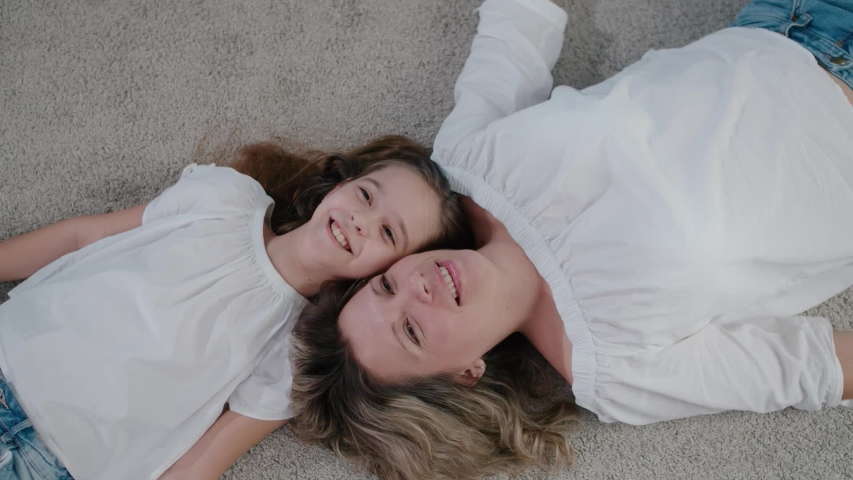 A beautiful mother and daughter lie on the floor inside the house and look up, smiling and having fun chatting. Top view Royalty-Free Stock Footage #1052963336