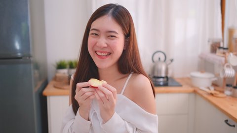 Beautiful woman slice red apple and bite to eat then smile and look to camera in kitchen with morning light. Concept of happy life to stay at home and social distance during covid virus pandemic.