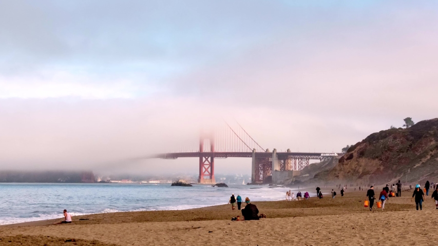 Time lapse of Golden Gate Bridge from Baker Beach - colorful morning light fog passing by as it slowly burns away from the sun - crowded beach tons of activity with lots of beach cleaning - 2019