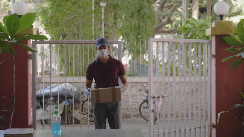 A delivery man or boy wearing a protective face mask delivers the box courier package on a building desk gate or doorstep during lockdown amid coronavirus or COVID19 epidemic or pandemic. Royalty-Free Stock Footage #1052973200