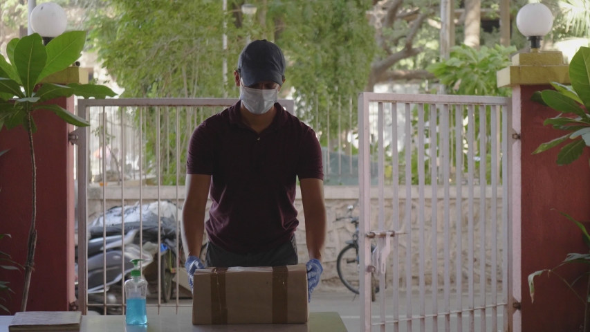 A delivery man or boy wearing a protective face mask delivers the box courier package on a building desk gate or doorstep during lockdown amid coronavirus or COVID19 epidemic or pandemic.