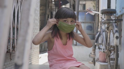 A close shot of cute young little girl child wearing or putting a face mask while sitting outside in the lane amid coronavirus or COVID19 epidemic or pandemic