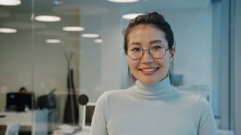 Portrait of attractive young Asian businesslady wearing glasses smiling standing in office alone near glass wall. Emotions, business and people concept.