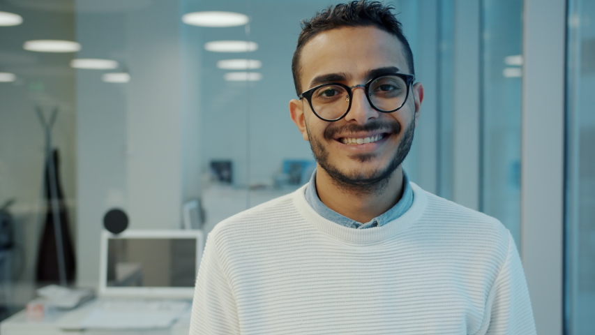 Portrait of handsome young Arab man office worker standing in workplace smiling looking at camera wearing glasses. Employees and workspace concept. Royalty-Free Stock Footage #1052973362