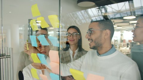 Arab businessman is using glassboard with sticky notes sharing ideas with coworkers solving business problems clapping hands. People and solutions concept.