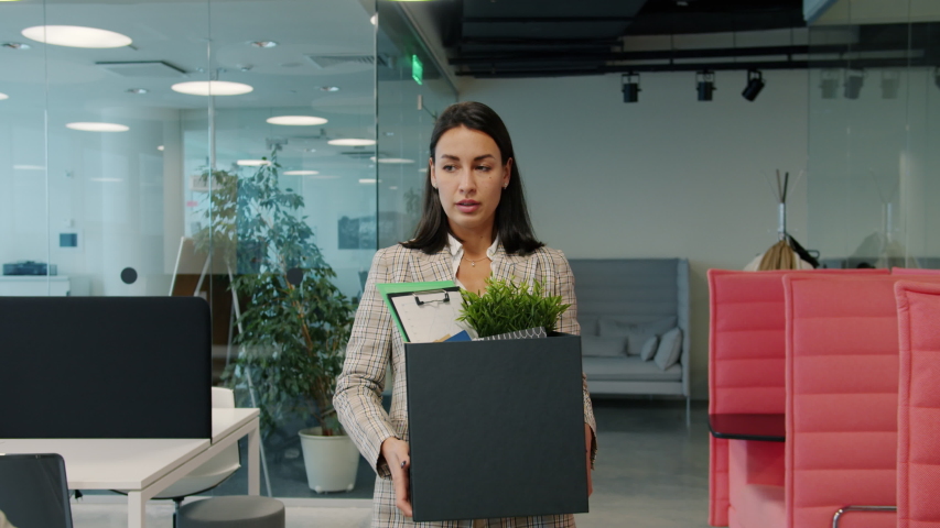 Sad young woman fired employee is leaving work with box of stuff walking in open space office while coworkers watching her with unhappy faces. Royalty-Free Stock Footage #1052973437
