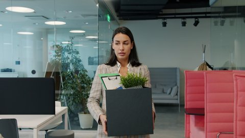 Sad young woman fired employee is leaving work with box of stuff walking in open space office while coworkers watching her with unhappy faces.