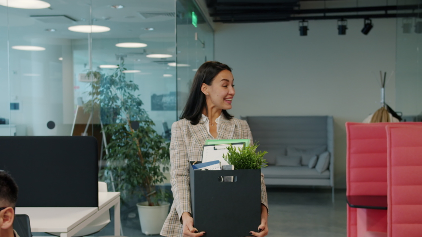 Happy young lady former employee is quitting job leaving office room with box of things walking and talking to ex-colleagues. People and employment concept. Royalty-Free Stock Footage #1052973446