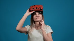 thoughtful young lady poses with modern orange virtual reality goggles on head standing on blue background slow motion