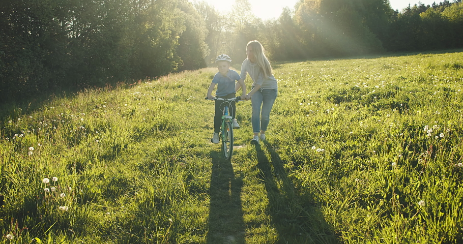 Mother teaching son to ride bicycle. Happy cute boy in helmet learn to riding a bike in park on green meadow in summer day at sunset time. Family weekend. 4K video Slow motion Royalty-Free Stock Footage #1052985236