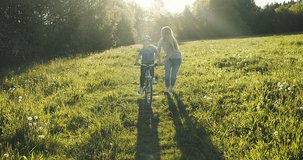 Mother teaching son to ride bicycle. Happy cute boy in helmet learn to riding a bike in park on green meadow in summer day at sunset time. Family weekend. 4K video Slow motion