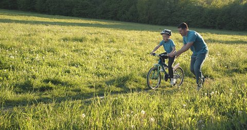 Father teaching son to ride bicycle. Happy cute boy in helmet learn to riding a bike in park on green meadow in summer day at sunset time. Family weekend. 4K video Slow motion