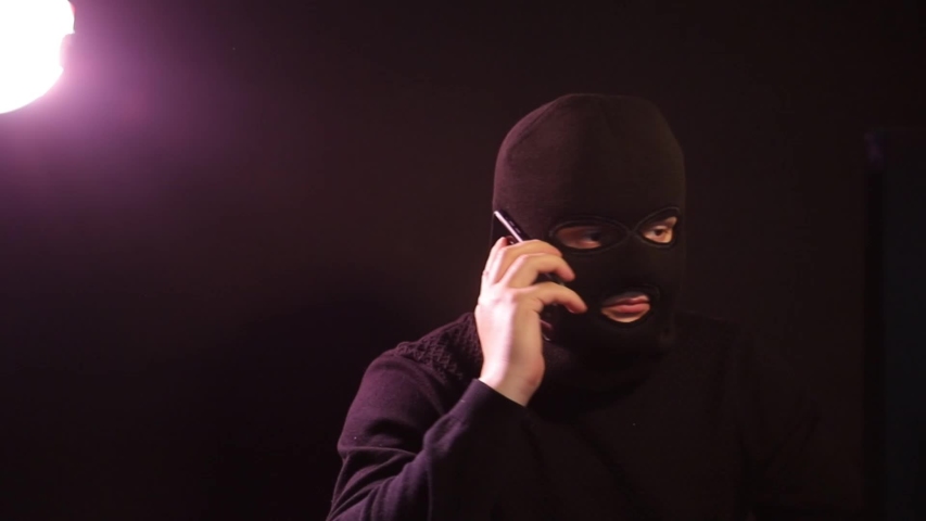 A man in a balaclava in a dark room talking on a smartphone. The criminal in a mask Royalty-Free Stock Footage #1052990321