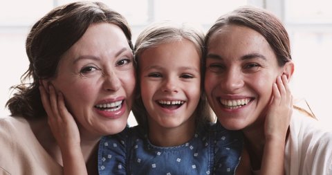 Close up portrait of happy beautiful three female generations family. Head shot little adorable preschool girl embracing smiling middle aged granny and pretty affectionate mother indoors.