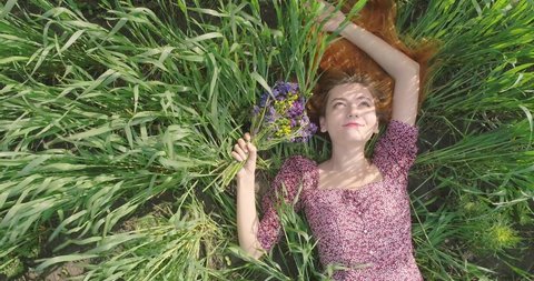 Video from the high of young red haired woman lying alone on huge green summer field in sunny weather. Aerial view of girl in purple dress relaxing on sunny grass in park. Summer holidays concept.