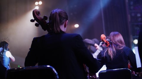 girls play the cello and violins at a concert, and in front of them, the director waves his wand