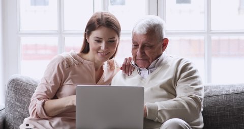 Smiling young attractive woman discussing internet shopping with older mature father, looking at computer screen. Happy grownup daughter showing technology application on laptop to old daddy at home.