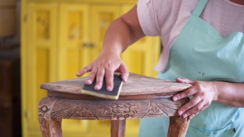 Close-up of craftswoman's hands using sandpaper and sanding antique ornate table in wood furniture workshop