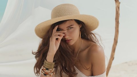Attractive brunette woman sitting on a deserted beach under the handmade tent. Gorgeous slim mixed race Asian Caucasian woman a white dress, hat, jewelry spending holidays on the sea shore. Tanned