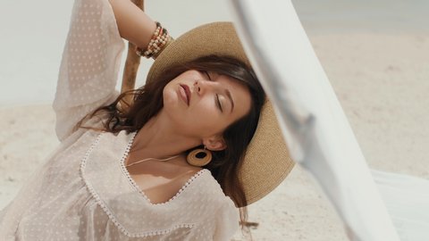 Seductive brunette woman sitting on a deserted beach under the handmade tent. Gorgeous slim mixed race Asian Caucasian woman a white dress, hat, jewelry spending holidays on the sea shore. Tanned