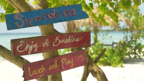 CLOSE UP, DOF: Vintage signs tell tourists to enjoy their day on the tropical beach. Colorful planks with positive sayings hang off a tree on a remote white sand beach in the picturesque Maldives.